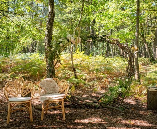 Clairière boisée aménagée pour une cérémonie de mariage, fauteuils en rotin et arche fleurie